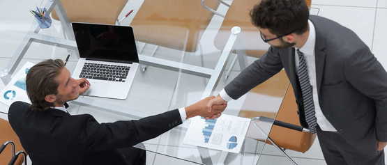 Two people shaking hands over a table with papers and a laptop.